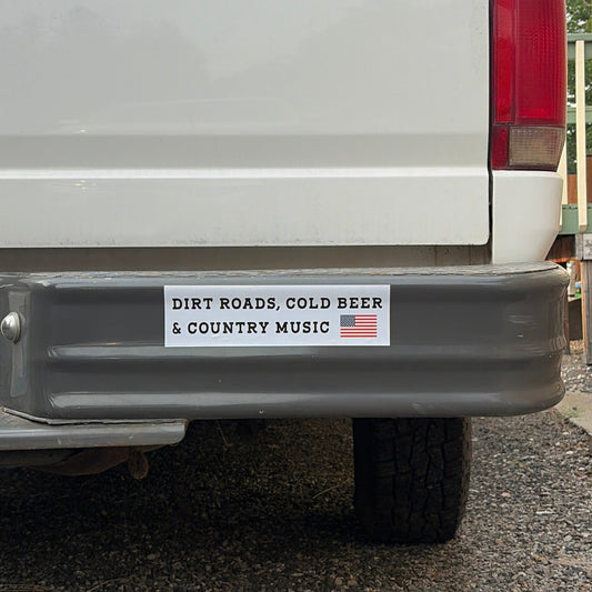 A rectangular bumper sticker on the rear bumper of a white pickup truck. The sticker has a white background with black text reading 'DIRT ROADS, COLD BEER & COUNTRY MUSIC' and a small American flag graphic on the right.
