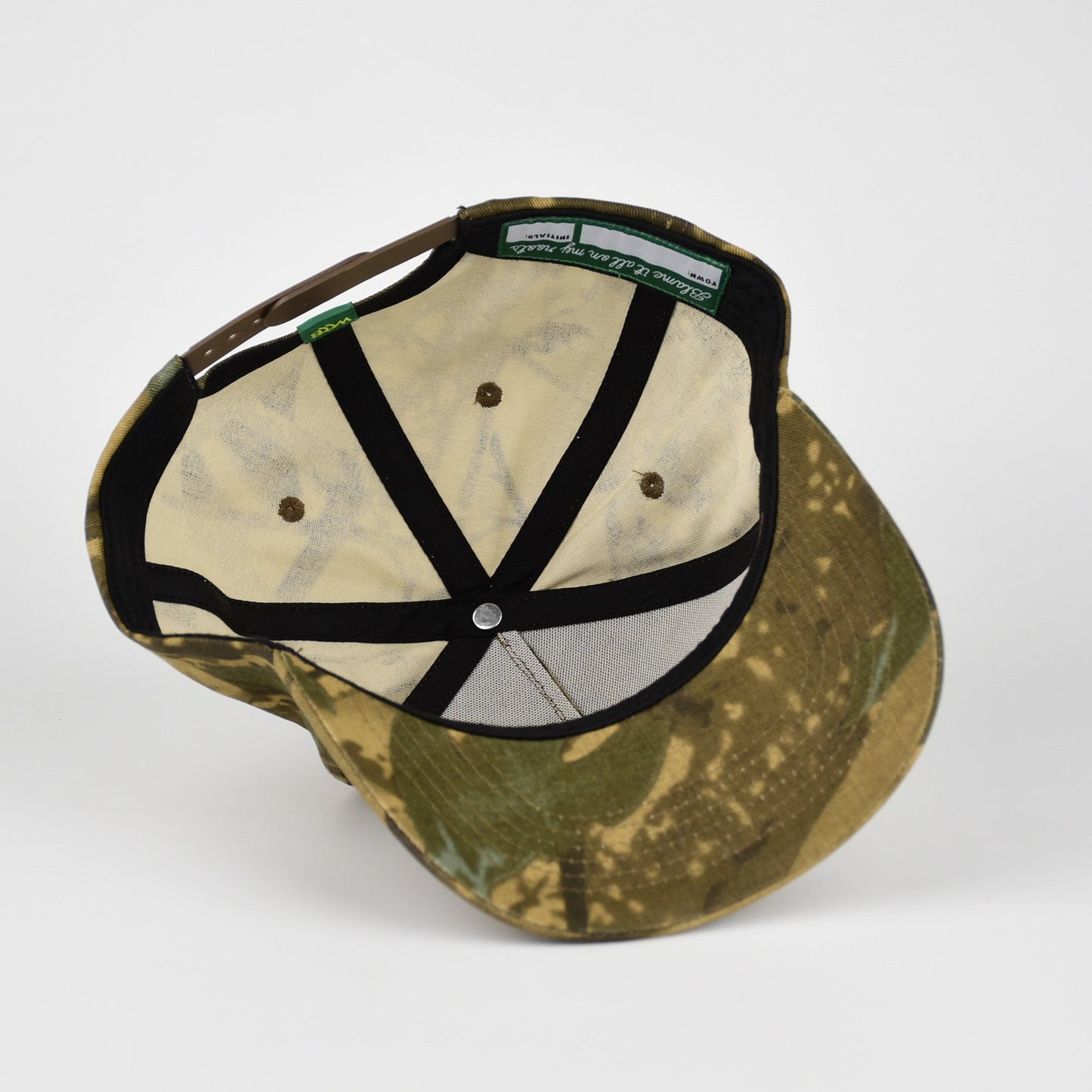 The underside of a camouflage baseball cap with black adjustable straps, photographed on a light gray background.
