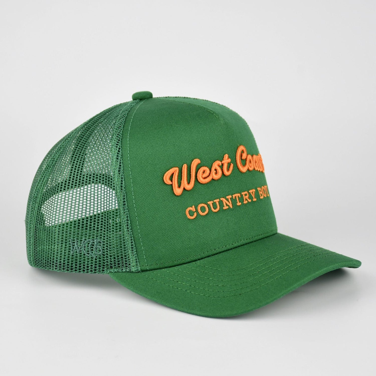 A green trucker-style baseball cap with a curved brim and mesh back, featuring orange embroidered text on the front that reads 'West Country COUNTRY BOY'.