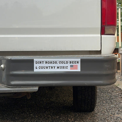 A rectangular bumper sticker on the rear bumper of a white pickup truck. The sticker has a white background with black text reading 'DIRT ROADS, COLD BEER & COUNTRY MUSIC' and a small American flag graphic on the right.