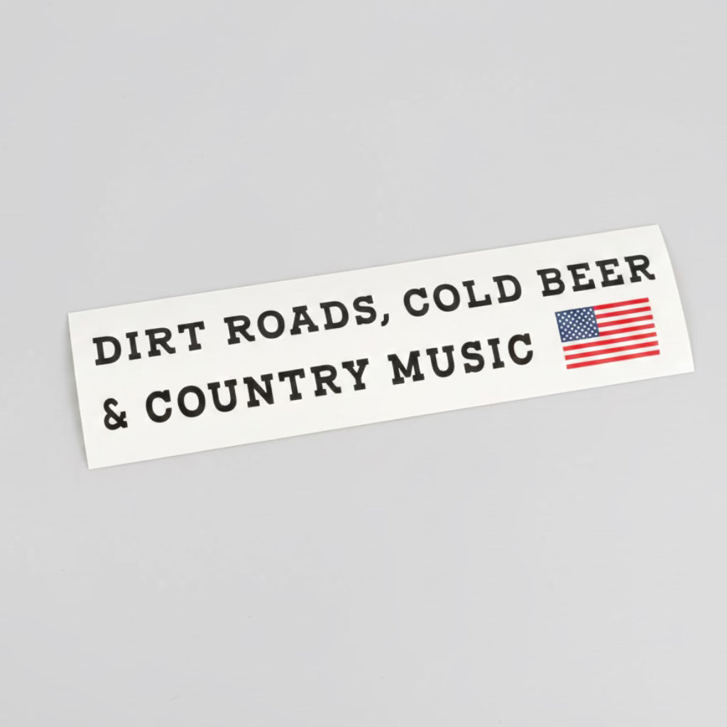 A white bumper sticker with black text reading 'DIRT ROADS, COLD BEER & COUNTRY MUSIC' and a small American flag graphic, placed on a dark tabletop.