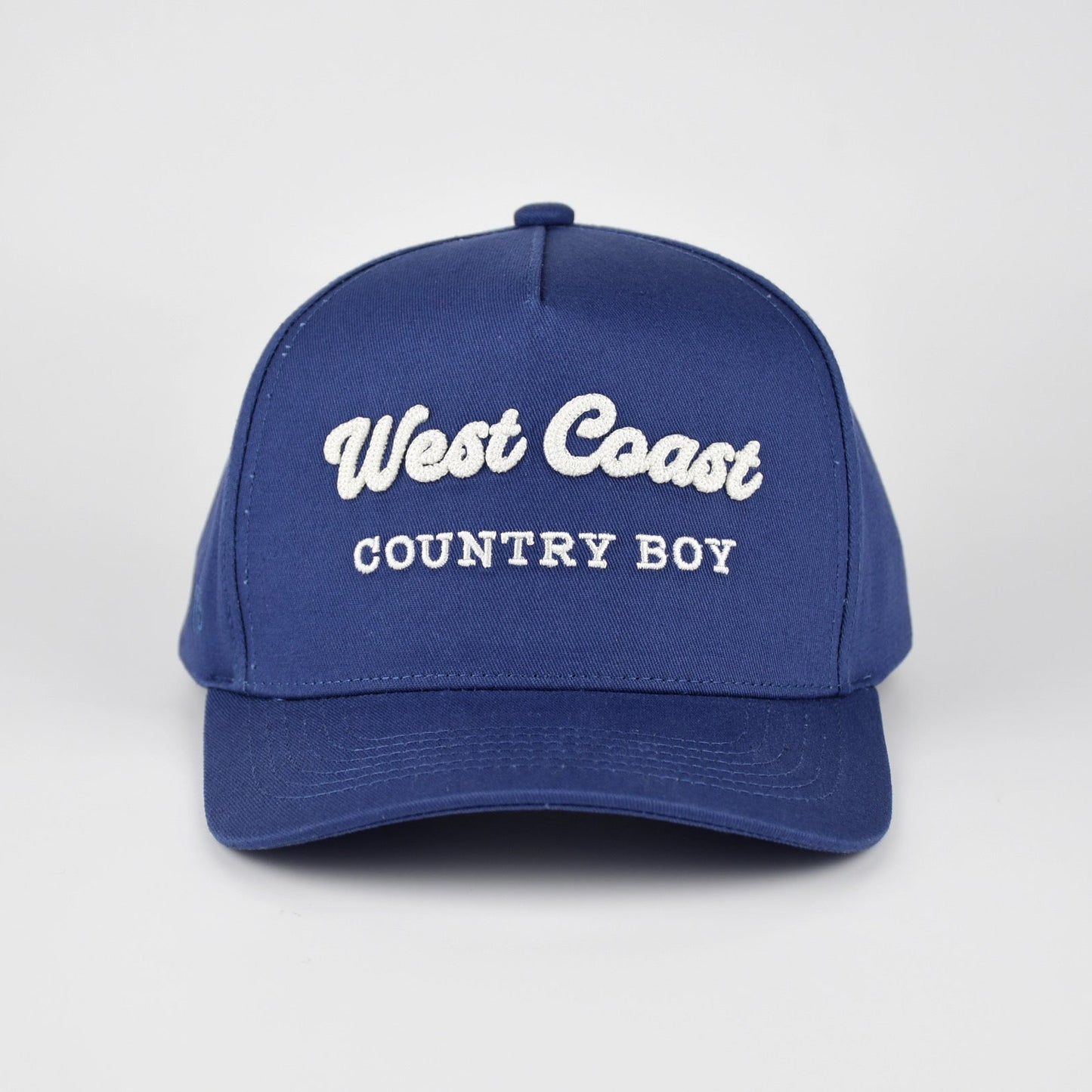 A royal blue baseball cap with 'West Coast COUNTRY BOY' embroidered in white on the front, set against a light gray studio background.