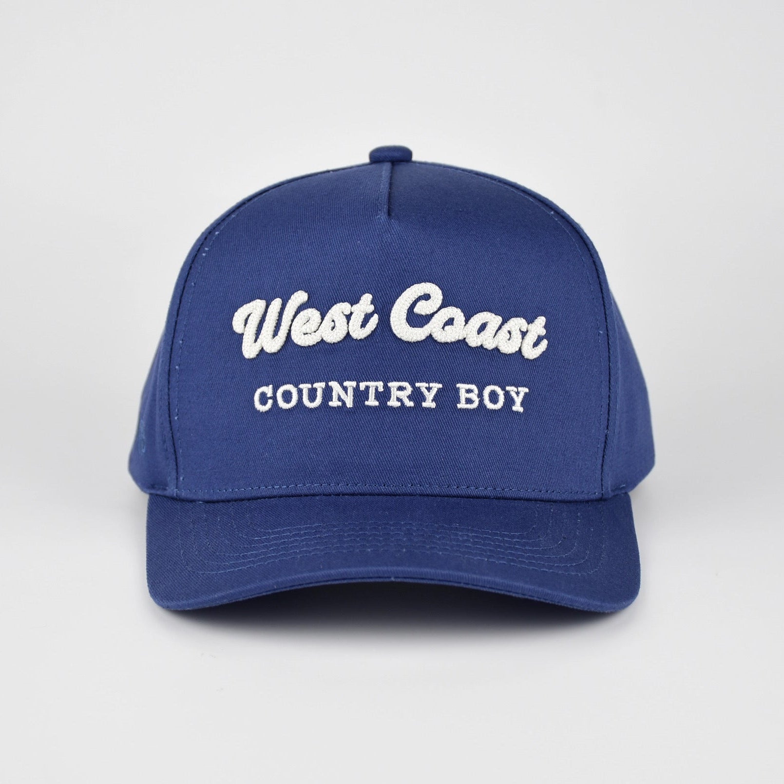 A royal blue baseball cap with 'West Coast COUNTRY BOY' embroidered in white on the front, set against a light gray studio background.