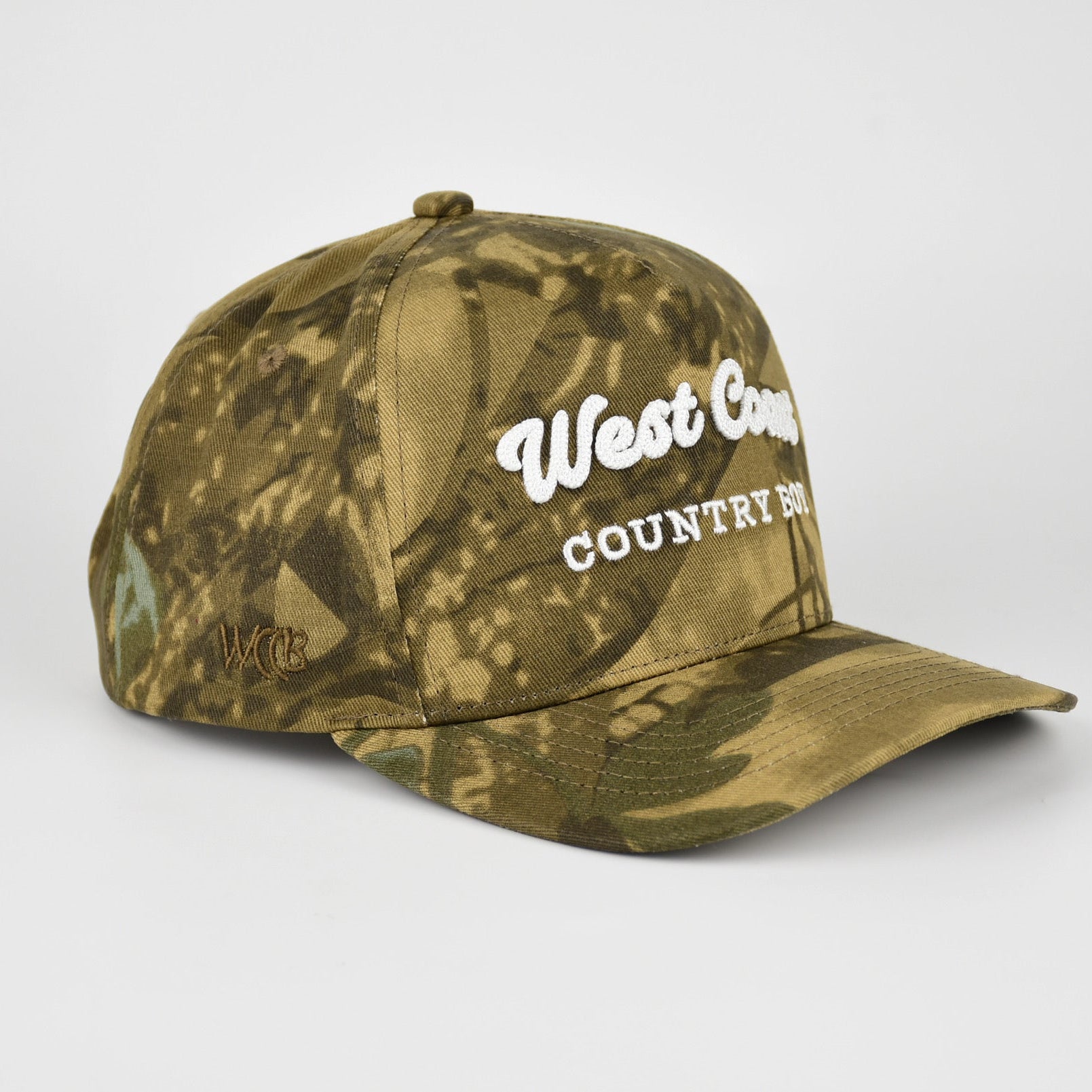 A structured baseball cap in a green and brown camouflage pattern with white embroidered text reading 'West Coast COUNTRY BOY', shown on a light gray studio background.