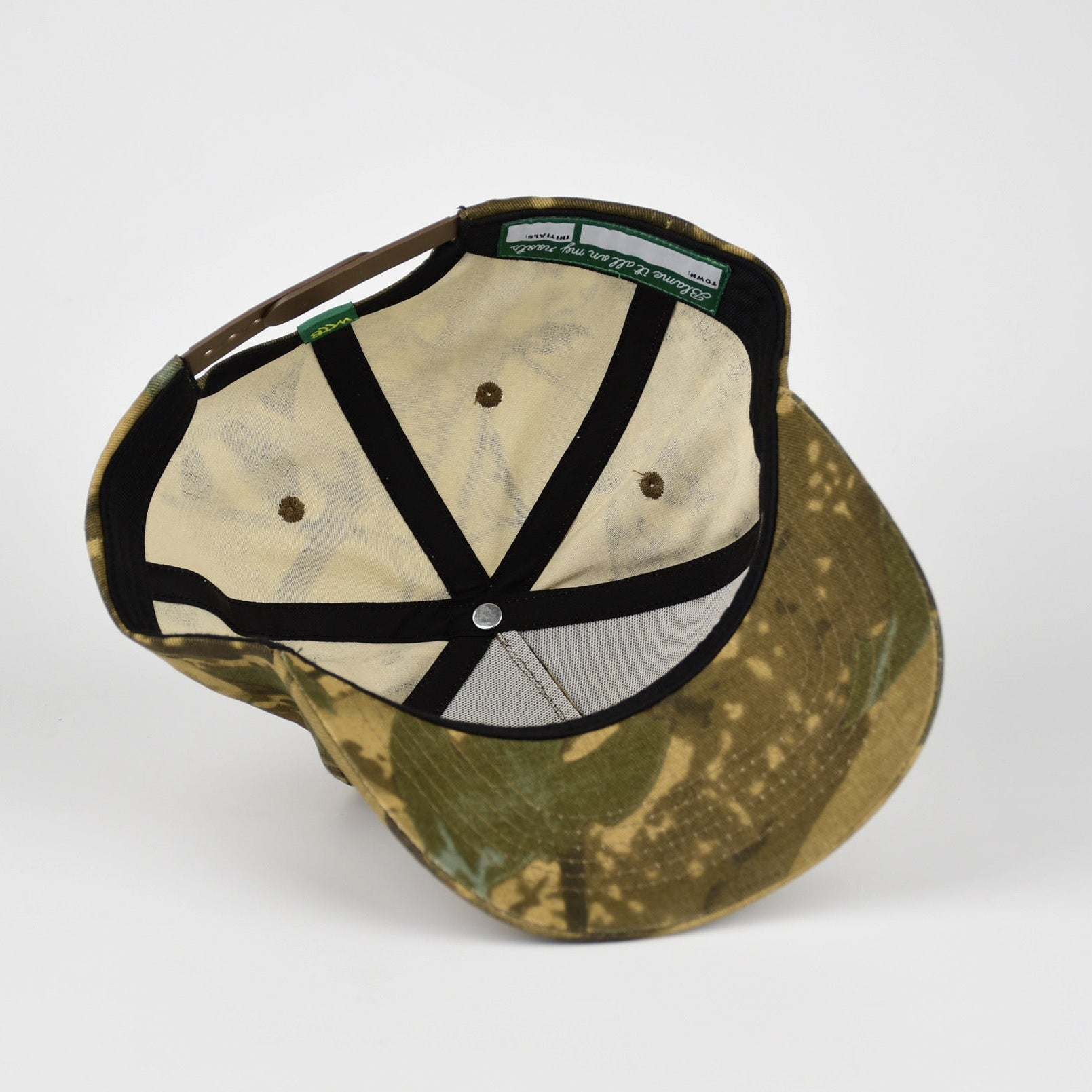 The underside of a camouflage baseball cap with black adjustable straps, photographed on a light gray background.