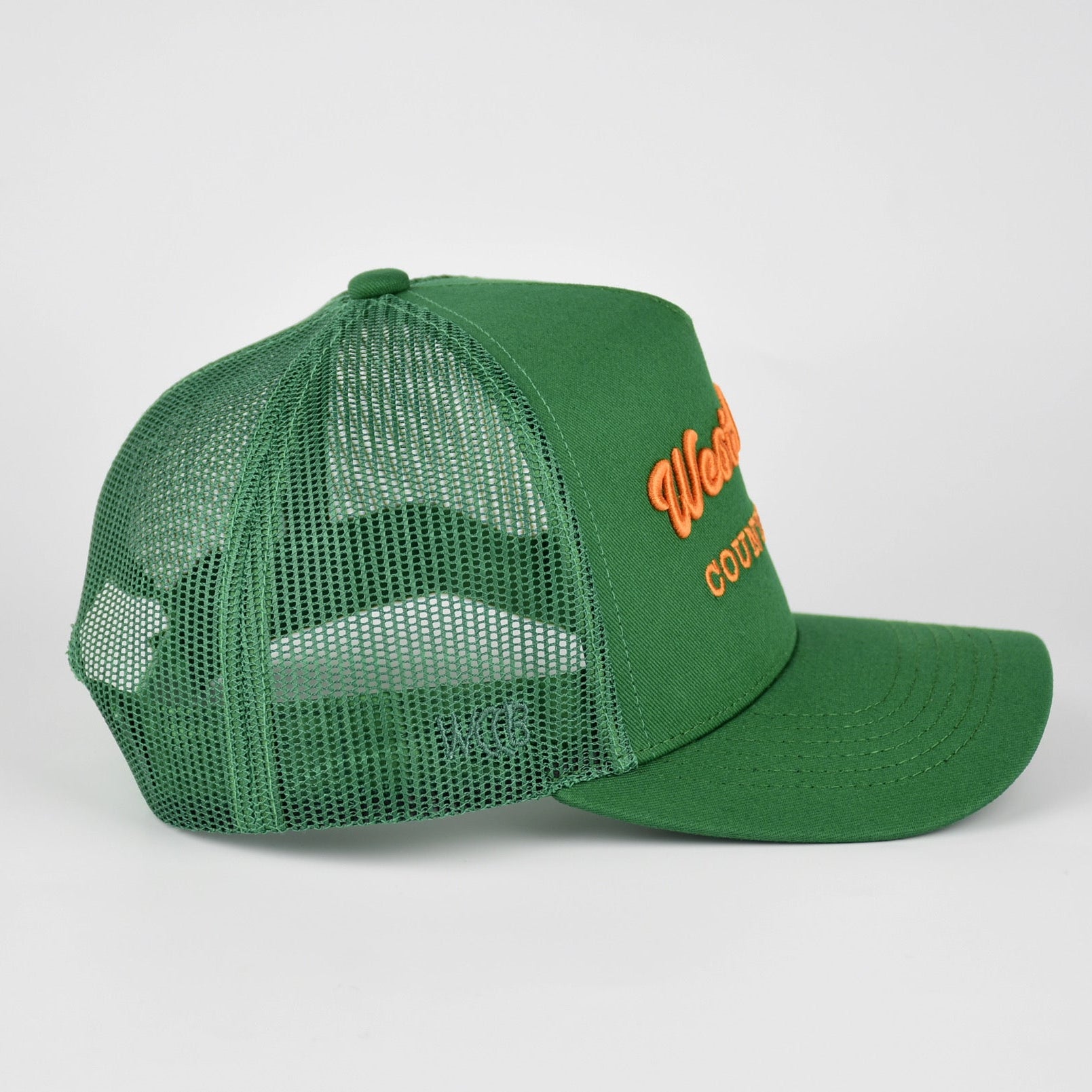 A green trucker-style baseball cap with a curved brim and mesh back, featuring orange embroidered text on the front panel and WCCB on the side shown against a light gray background.