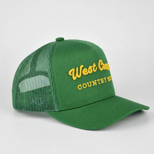 A green trucker-style baseball cap with a curved brim and mesh back, featuring yellow embroidered text on the front that reads 'West Country COUNTRY BOY.